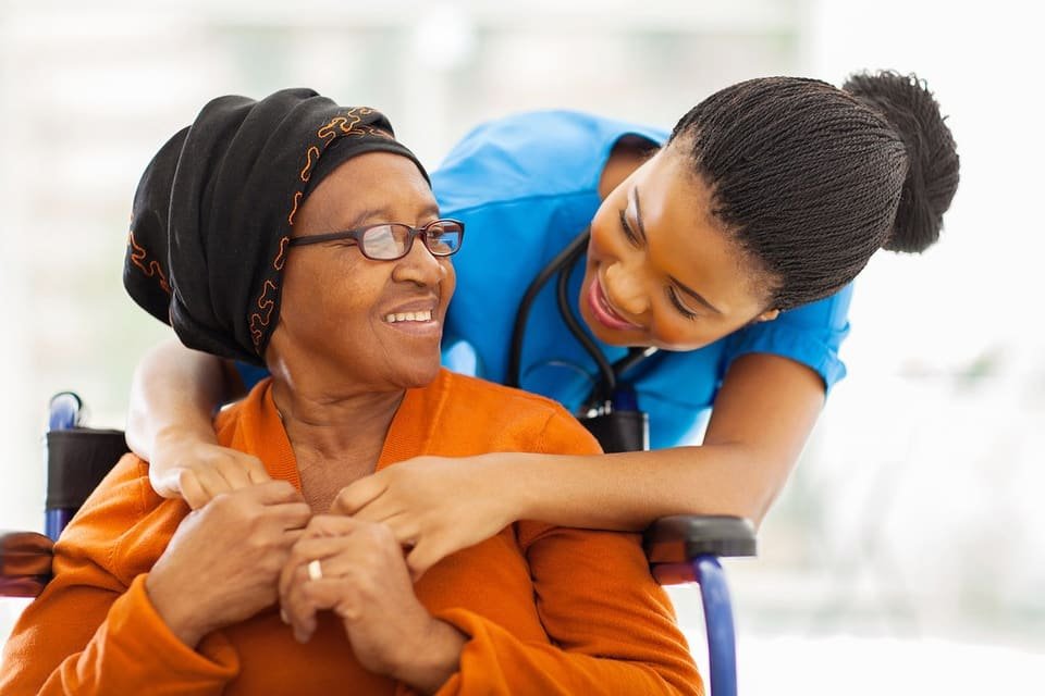 A smiling caregiver hugs an older woman in an orange top from behind as they share a warm moment