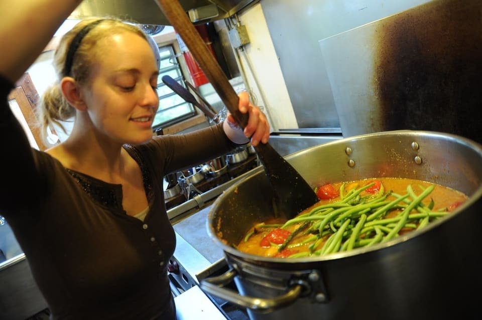 A smiling woman stirs a large pot of green bean soup in a busy kitchen