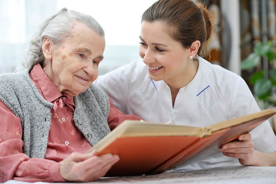 Placeholder portrait: a caregiver and an older woman reading a book together at the kitchen table