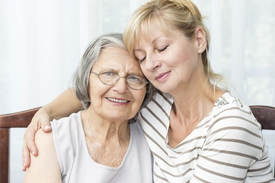 An adult daughter hugs her smiling mother on the sofa at home