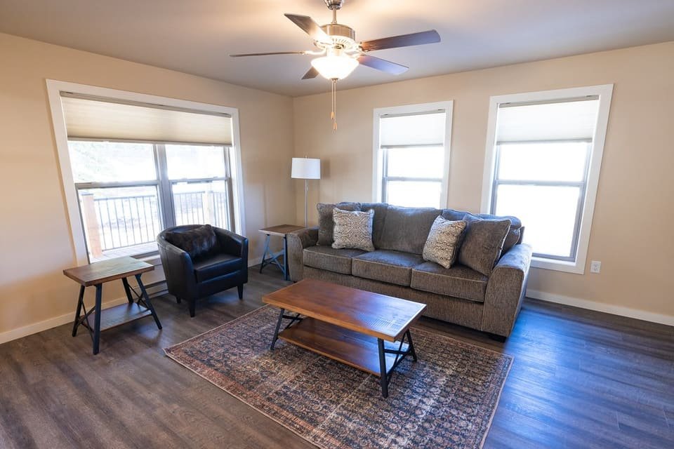 A bright, furnished living room with a grey sofa, wooden coffee table, rug and three large windows